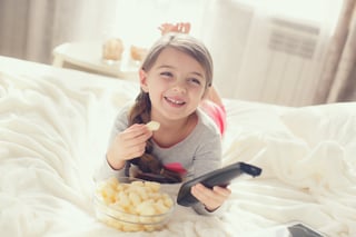 The cheerful girl eats popcorn and changes channels of the TV lying in a bed